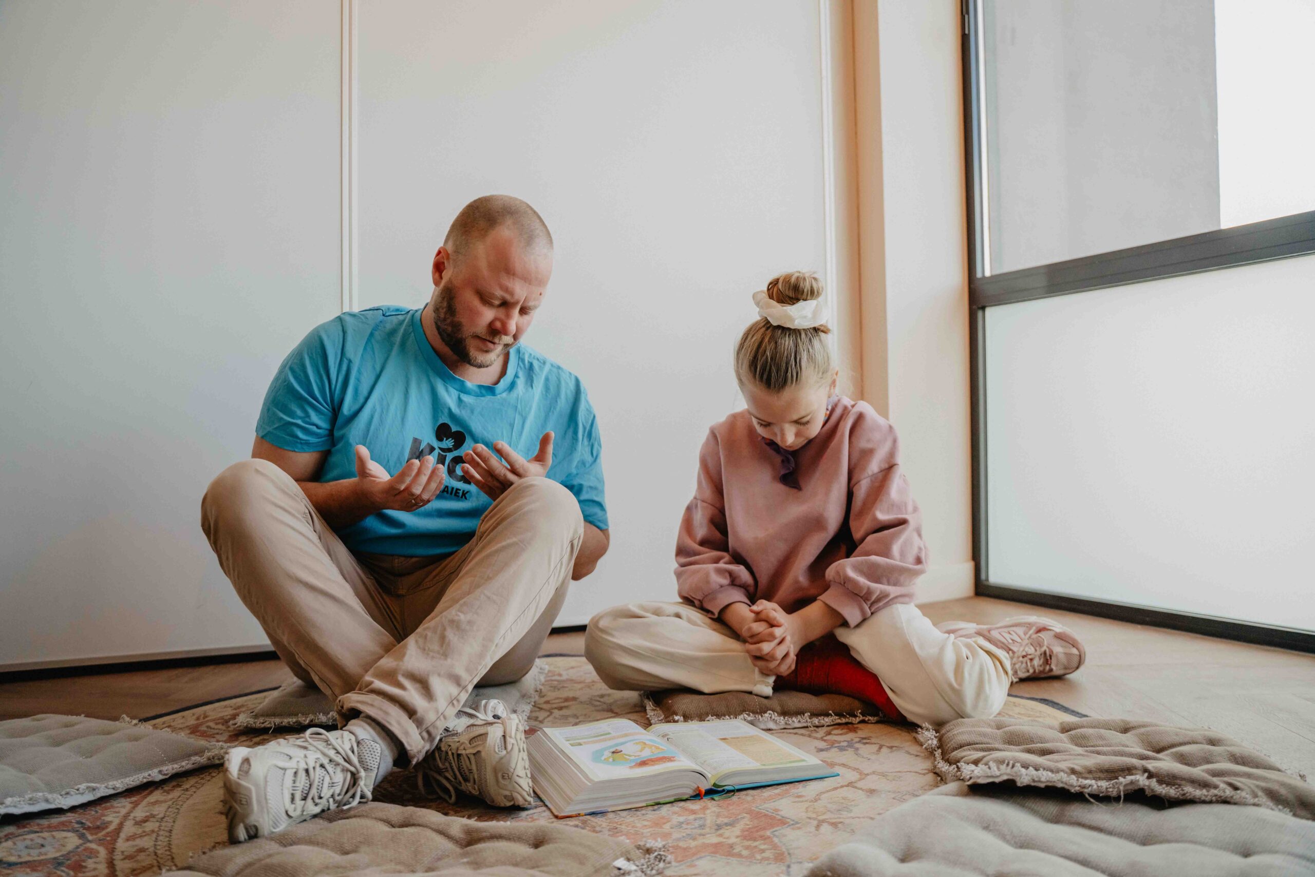 Vrijwilliger en meisje zitten samen op de grond bij een open kinderbijbel