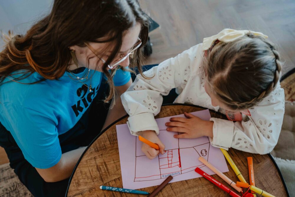 Vrijwilliger in Kids-shirt helpt een meisje met tekenen aan tafel