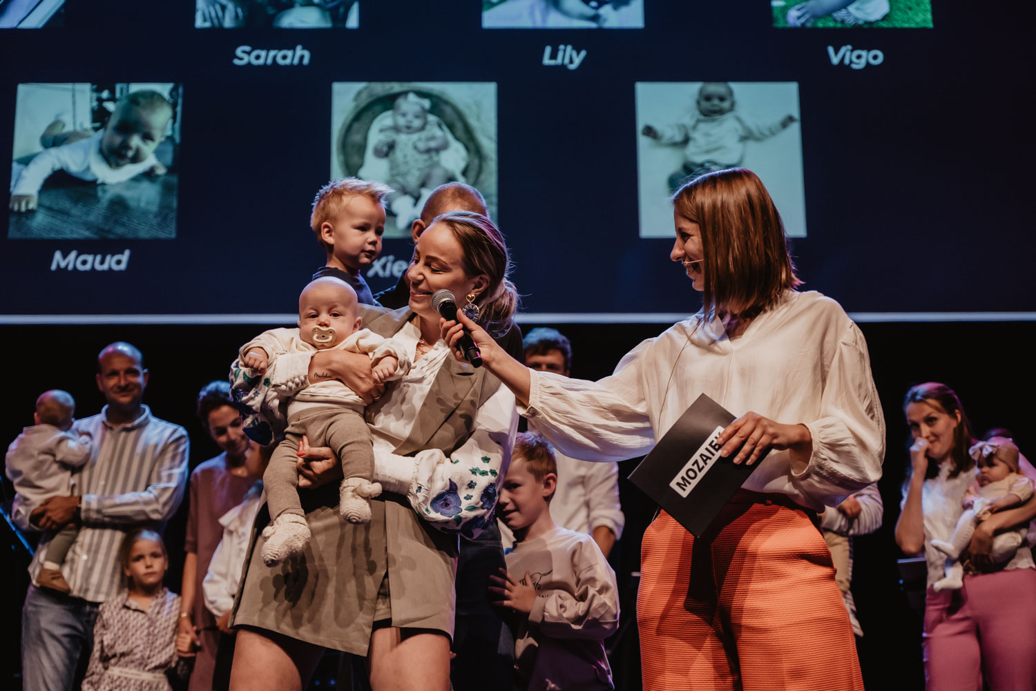 Gezinnen met hun babys op het podium, met de namen van de kinderen op het scherm