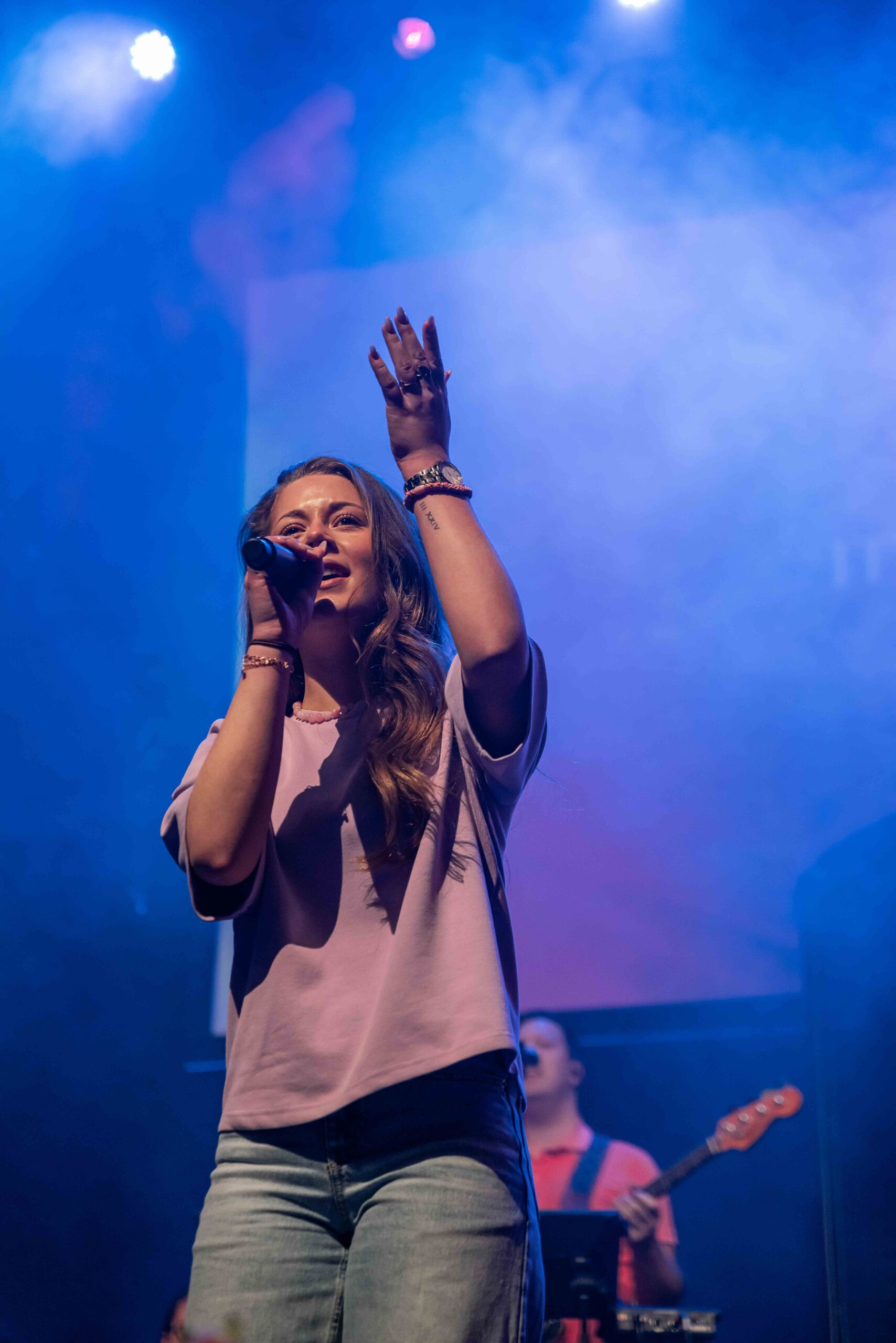 Zangeres zingt met opgeheven hand in blauw licht, met de bassist op de achtergrond