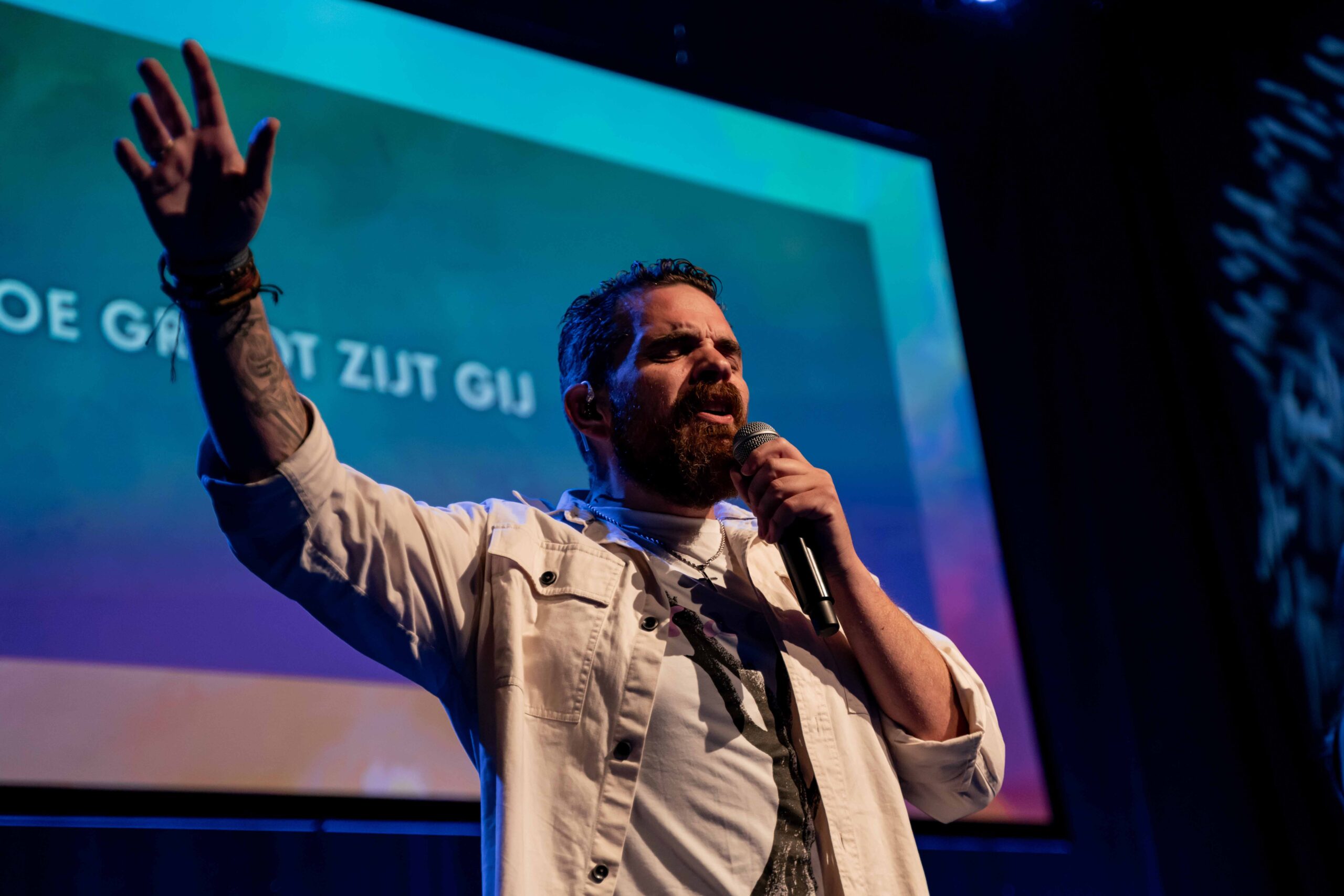 Worshipleider zingt met opgeheven hand voor het scherm met de songtekst Hoe groot zijt Gij